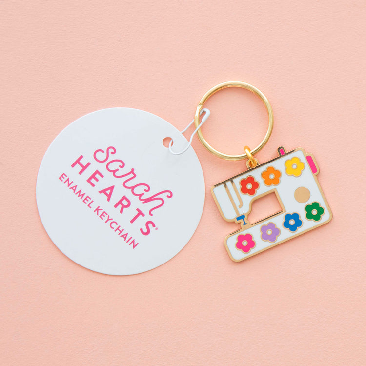 Enamel keychain with sewing machine design and 'Sarah Hearts' branding on a pink background